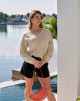 Woman standing by a pool holding a red handbag, wearing a beige sweater and black shorts, with a scenic background.