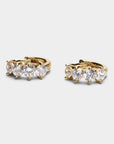 A pair of gold huggie hoop earrings with five round-cut clear gemstones in a row, set against a light gray background.