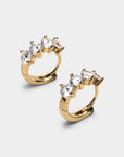 A pair of gold huggie hoop earrings with a curved top set with three clear marquise-cut gemstones, shown on a light gray background.
