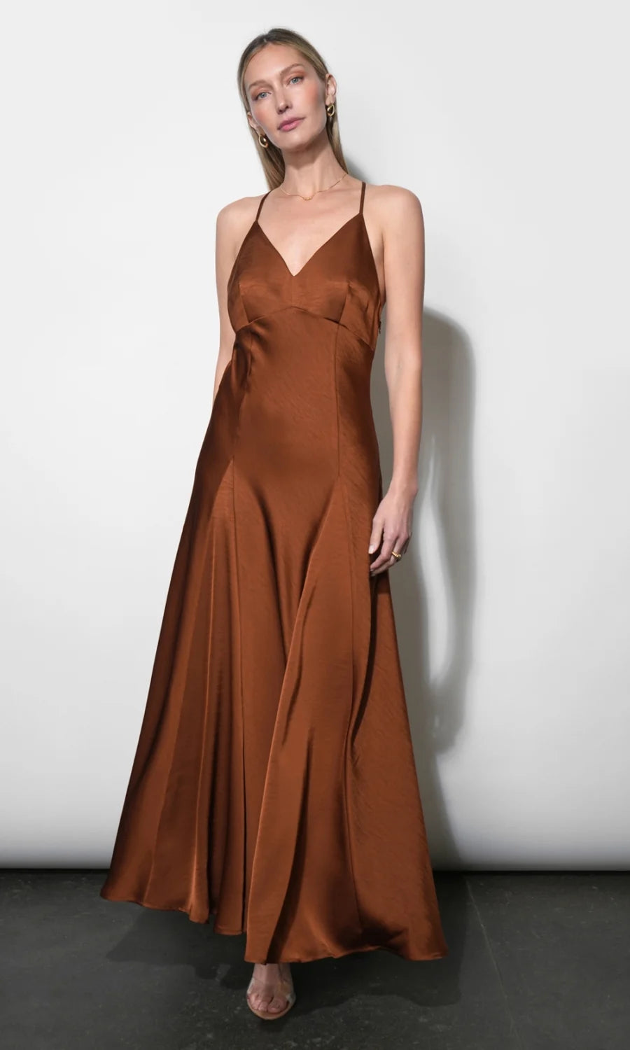 A woman modeling a floor-length satin slip dress in a warm rust brown color, standing against a plain light gray wall.