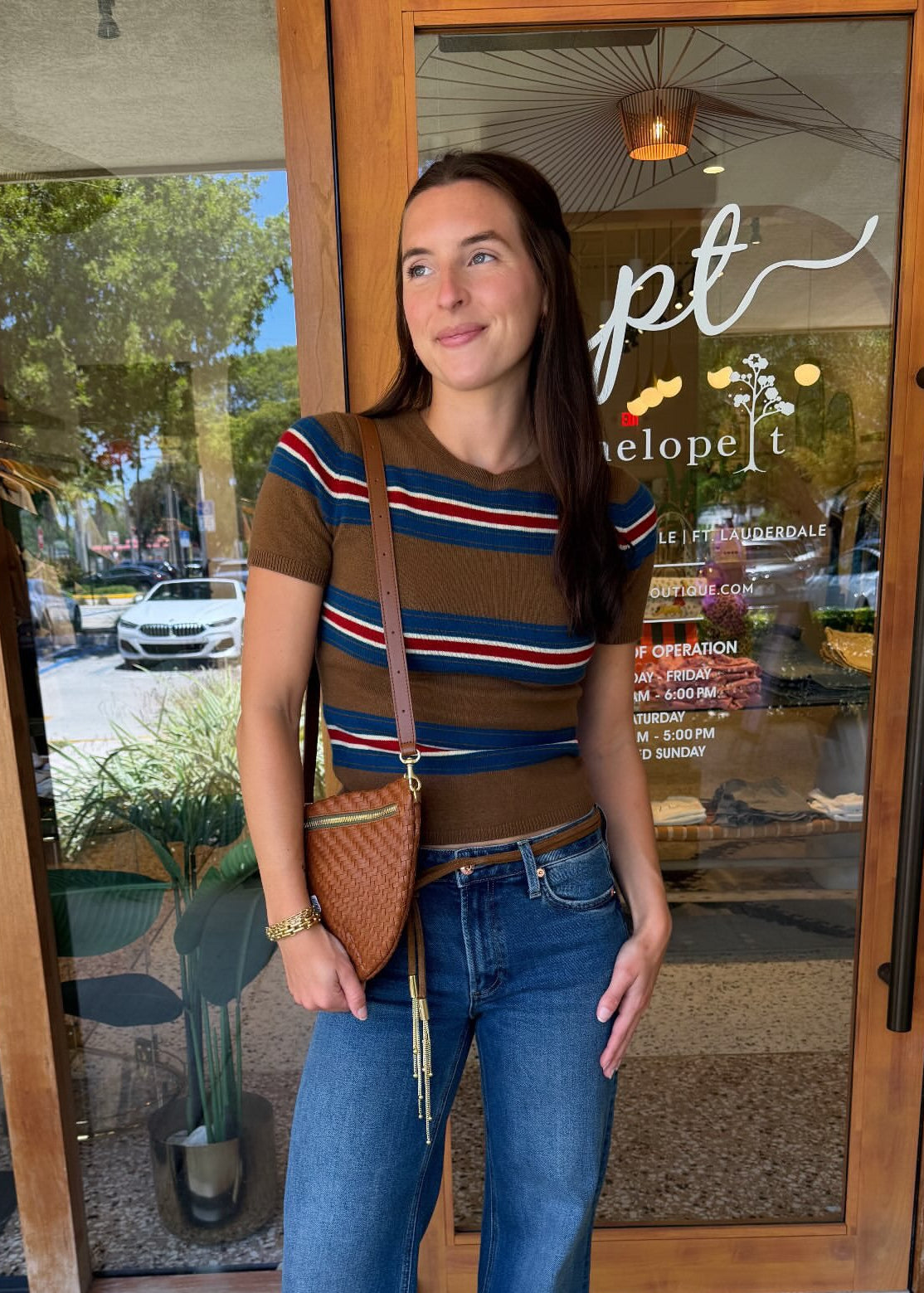 A woman stands on a sidewalk in front of a storefront, wearing a brown short-sleeve knit top with red, blue, and white horizontal stripes, blue wide-leg jeans, and carrying a brown woven crossbody bag. A white label at the bottom right reads 'New Staud Fall'.
