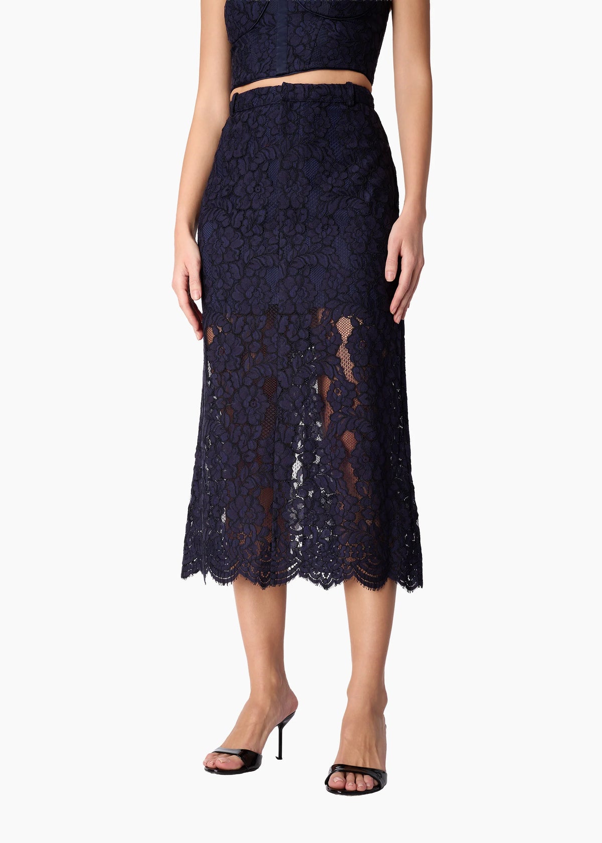 A model wearing a dark navy blue lace midi skirt with a scalloped hem, paired with a matching crop top and black heeled sandals, shown against a plain white background.