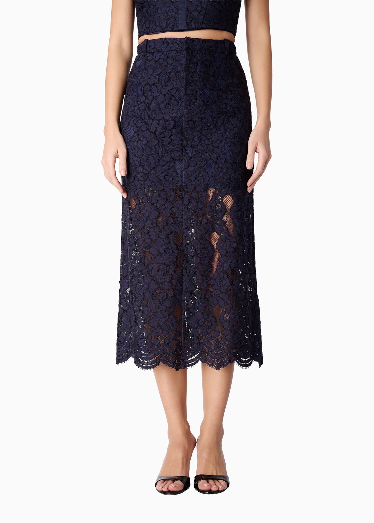 A model wearing a dark navy blue lace midi skirt with a scalloped hem, paired with a matching crop top and black sandals, shown against a plain white background.