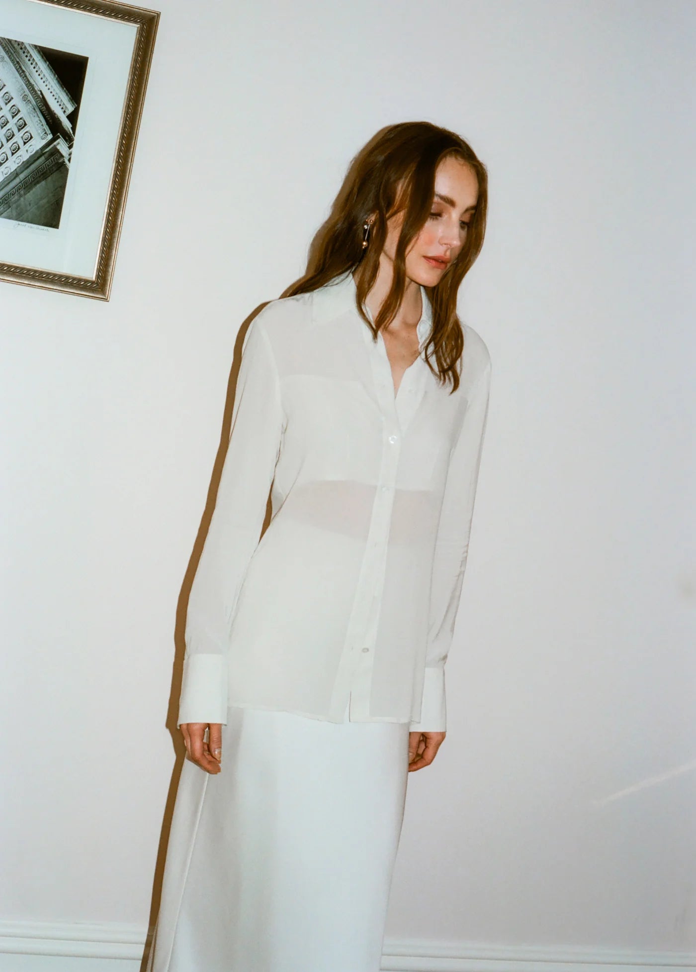 Woman wearing a white blouse and skirt standing against a plain wall.