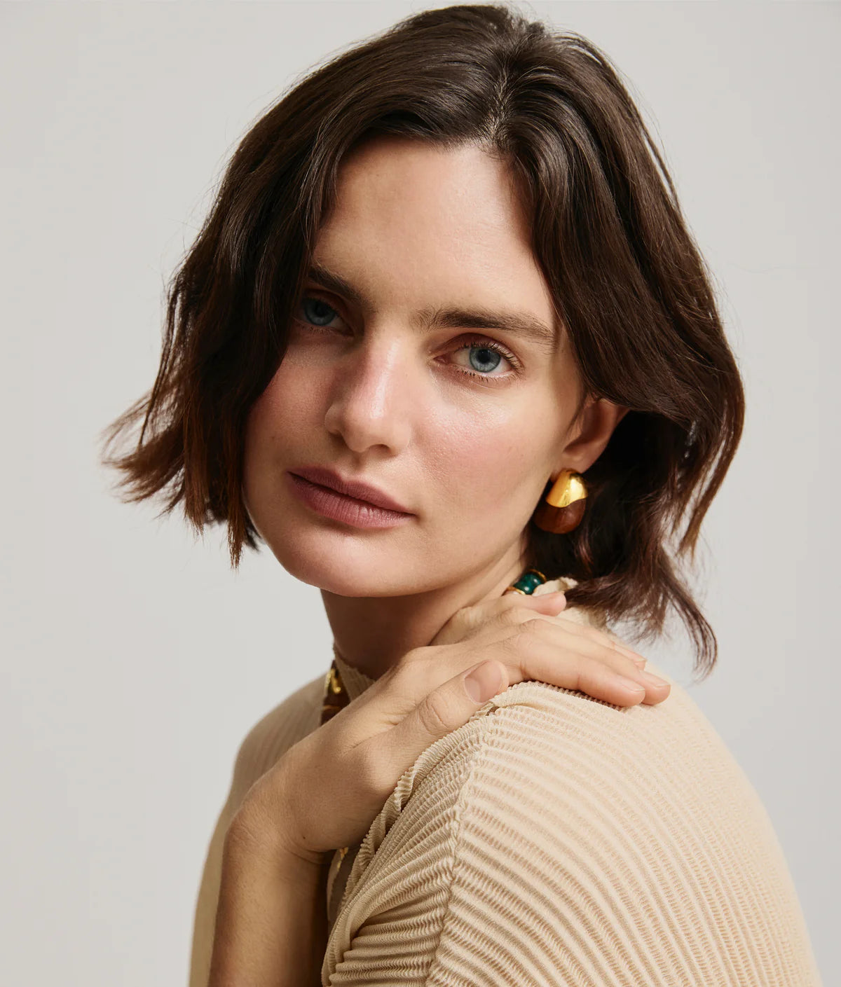 Woman with short brown hair wearing wood and gold earrings and a beige sweater against a light background
