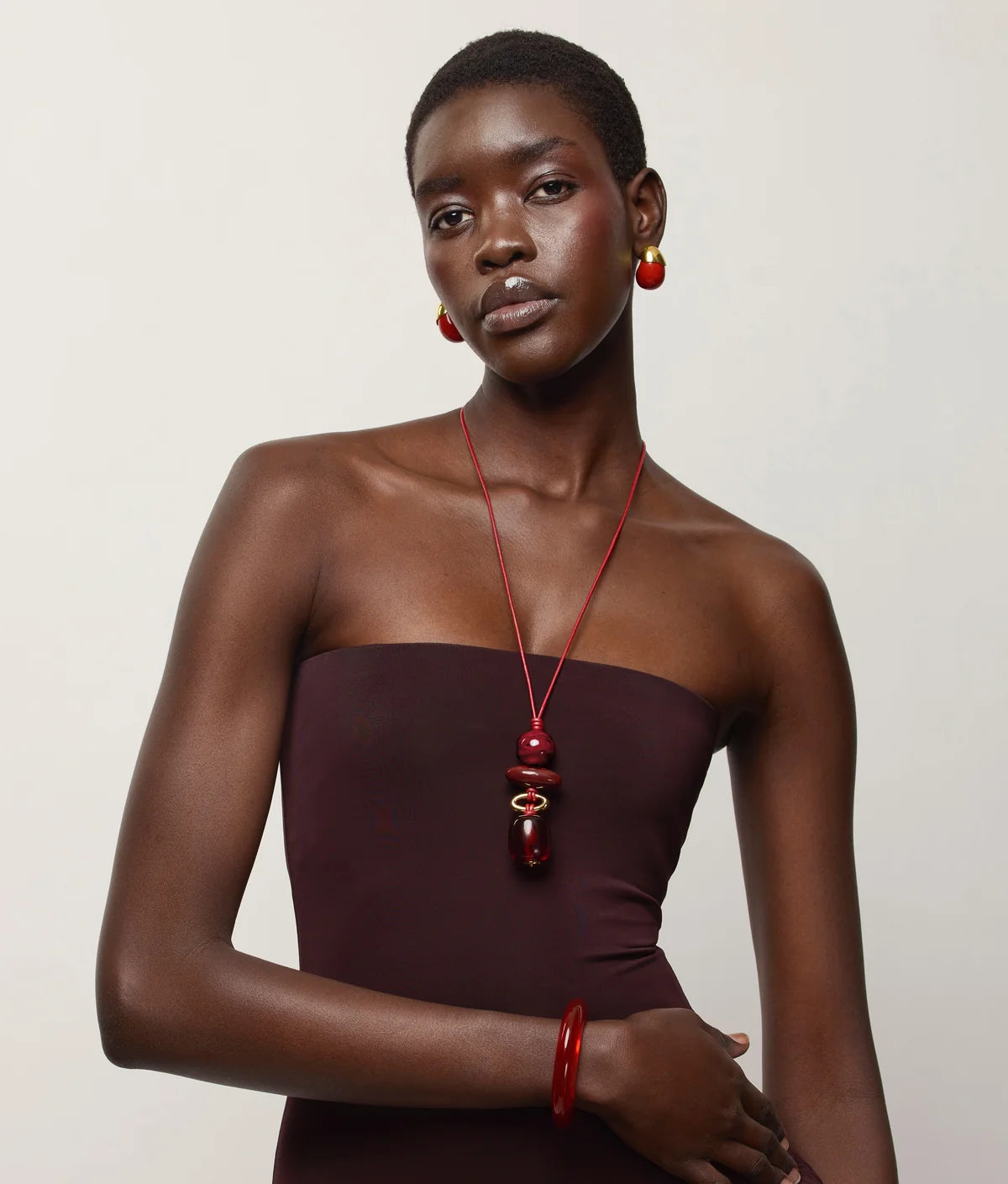 A model wearing a dark plum strapless dress, a red beaded necklace, a red bangle, and red hoop earrings, posed against a light gray studio background.