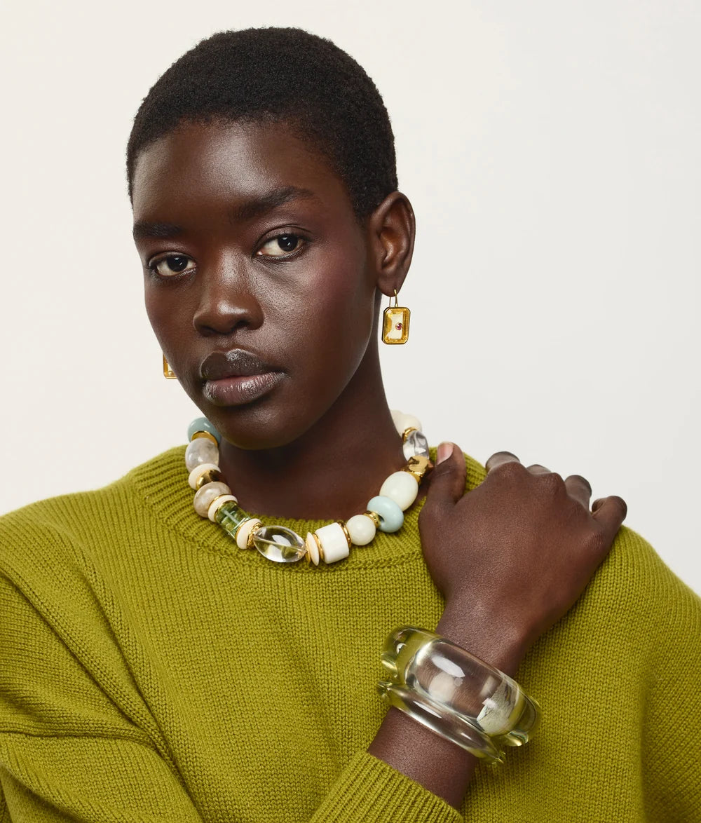 A model with short dark hair is shown from the chest up, wearing a textured olive green knit sweater, a chunky beaded necklace with white, cream, and light green beads, gold-toned earrings, and a clear acrylic bangle. The background is a clean, solid off-white studio backdrop.