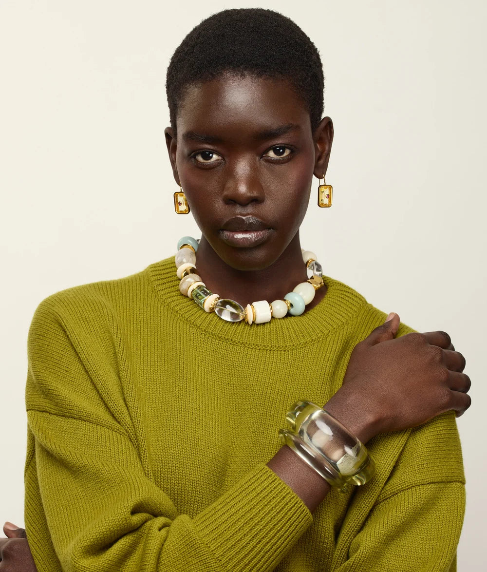 A model with short dark hair is wearing a textured olive green knit sweater, a chunky beaded necklace, gold rectangular earrings, and a wide clear resin cuff bracelet, posed against a solid off-white background.