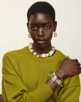 A model with short dark hair is wearing a textured olive green knit sweater, a chunky beaded necklace, gold rectangular earrings, and a wide clear resin cuff bracelet, posed against a solid off-white background.