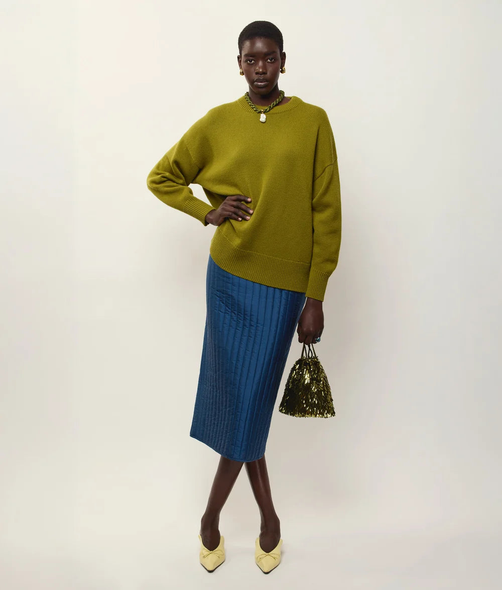 A model stands against a plain light background wearing an olive green knit sweater, a blue pleated midi skirt, and holding a small green sequined handbag.