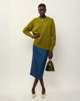 A model stands against a plain light background wearing an olive green knit sweater, a blue pleated midi skirt, and holding a small green sequined handbag.