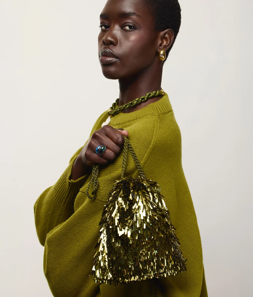 A model wearing a textured olive green sweater and matching green braided accessories, holding a gold sequined fringe handbag against a plain light background.