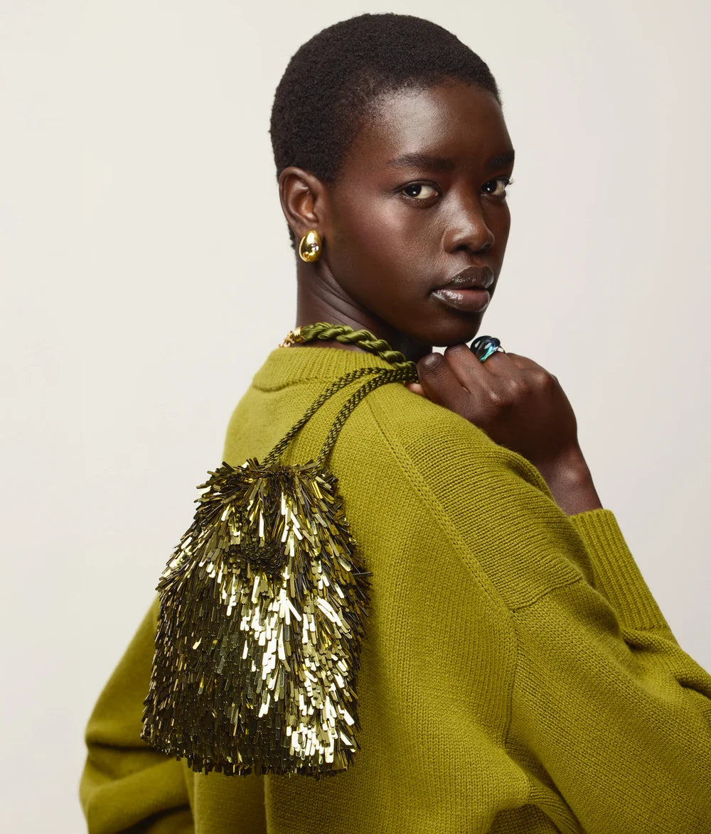 A model with short dark hair is shown from the back and side, wearing a textured olive green knit sweater and carrying a gold sequined shoulder bag against a plain light beige background.