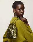 A model with short dark hair is shown from the back and side, wearing a textured olive green knit sweater and carrying a gold sequined shoulder bag against a plain light beige background.