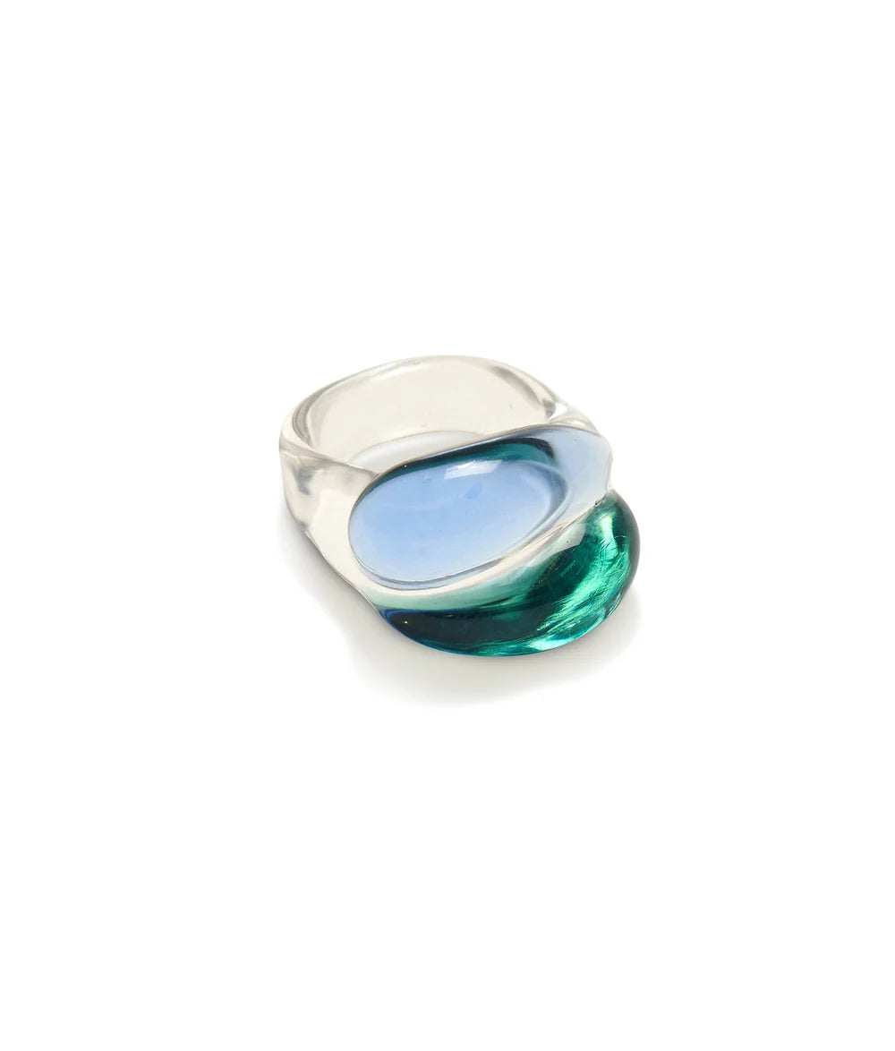 A modern sculptural ring featuring a wide, polished silver band with a large, organic-shaped inlay of translucent blue and green glass, set against a clean white background.