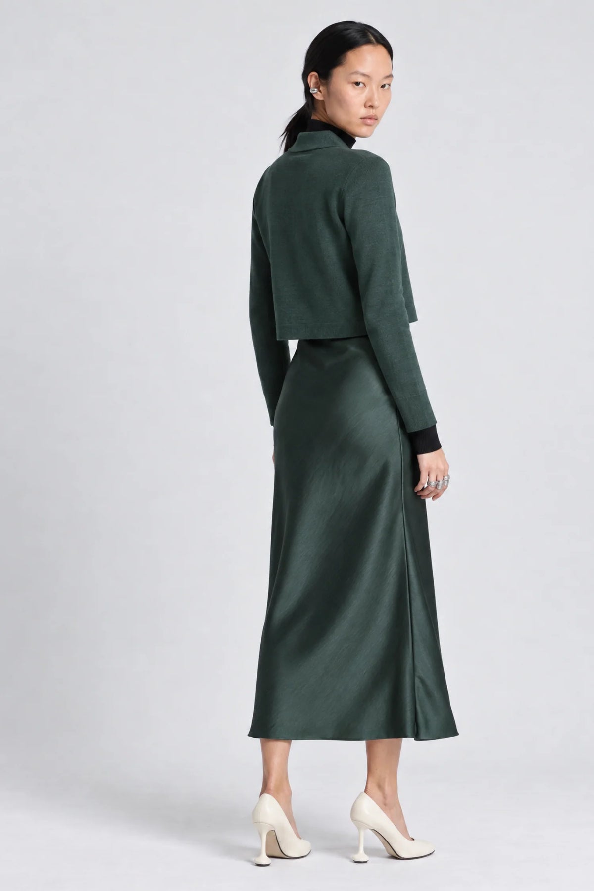A model is shown from the back wearing a dark forest green two-piece set consisting of a cropped long-sleeve sweater and a matching midi skirt, paired with cream-colored high-heeled pumps against a light gray studio background.