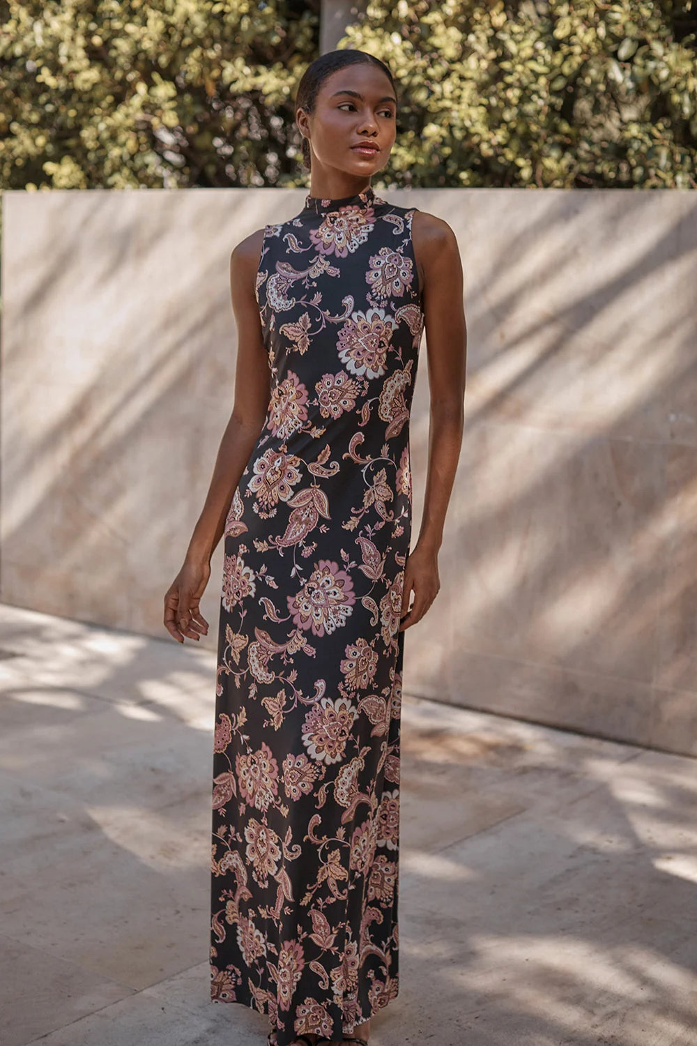 A woman wearing a sleeveless, high-neck, floor-length black dress with a pink and beige floral paisley pattern, standing outdoors against a beige stone wall with dappled sunlight.