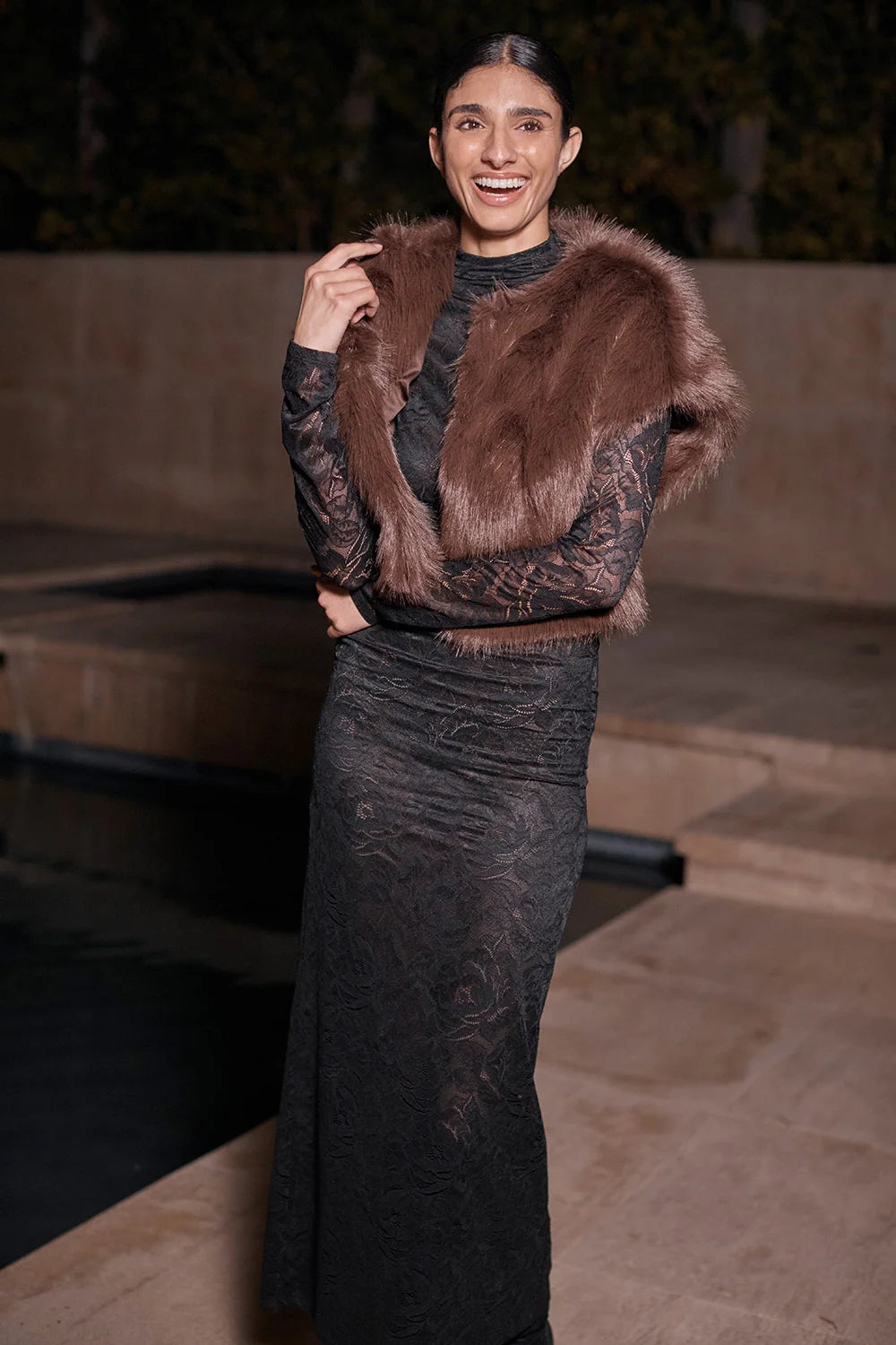 A woman wearing a dark lace long dress with a brown fur vest, standing outdoors at night near a stone poolside setting.