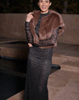 A woman wearing a dark lace long dress with a brown fur vest, standing outdoors at night near a stone poolside setting.