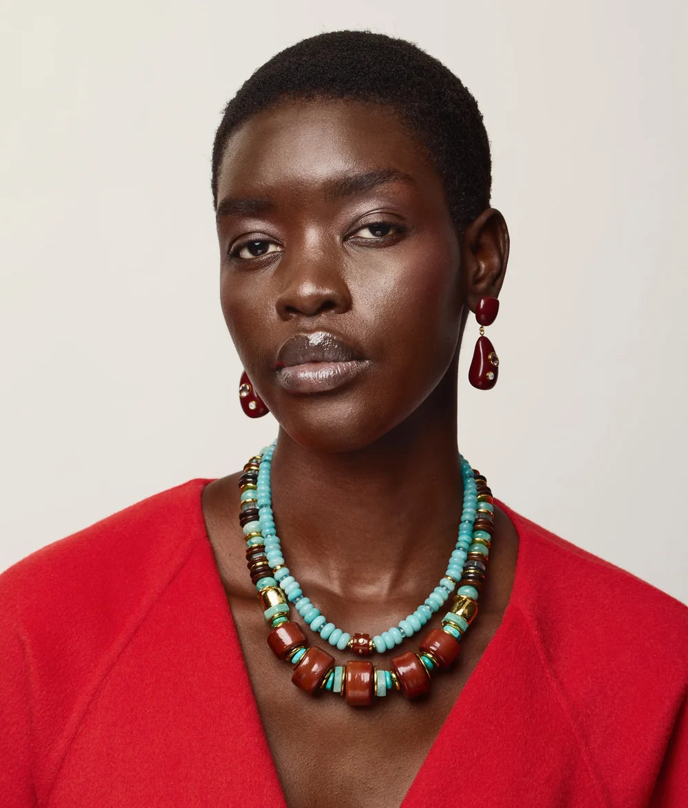 A close-up portrait of a model wearing a multi-strand turquoise and brown beaded necklace with matching drop earrings, paired with a bright red top against a light beige background.