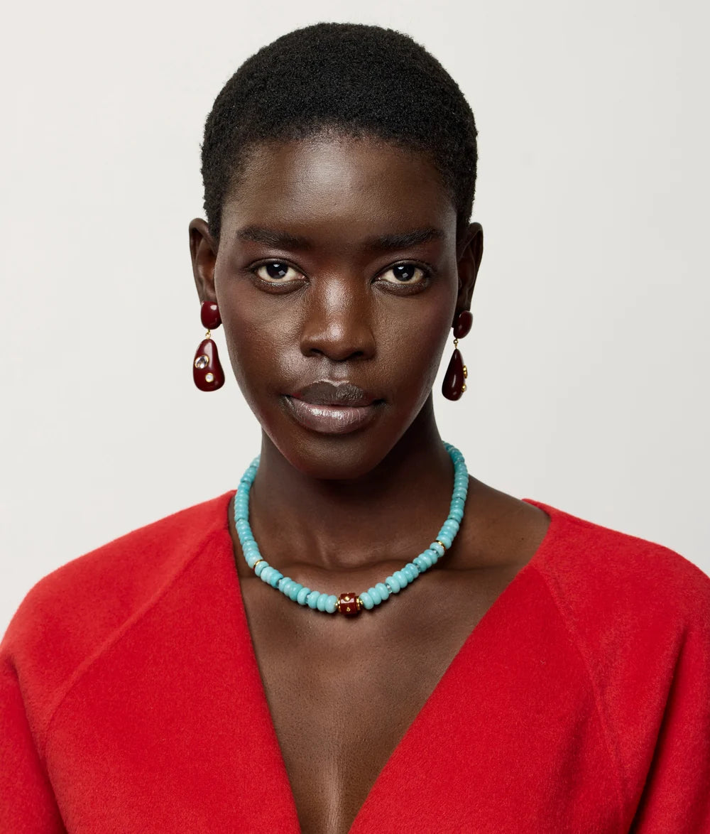A close-up portrait of a model wearing a turquoise beaded necklace with a central gold accent, matching teardrop earrings, and a bright red top against a light gray background.