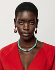 A close-up portrait of a model wearing a turquoise beaded necklace with a central gold accent, matching teardrop earrings, and a bright red top against a light gray background.
