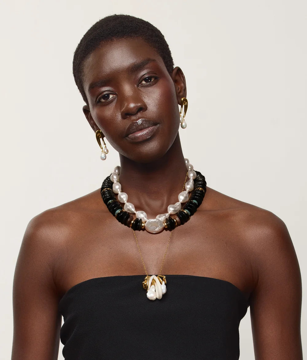 A model wearing a multi-strand necklace with white pearls, black beads, and gold-toned accents, along with matching gold hoop earrings and a gold pendant necklace, styled with a black strapless top against a light beige background.