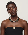 A model wearing a multi-strand necklace with white pearls, black beads, and gold-toned accents, along with matching gold hoop earrings and a gold pendant necklace, styled with a black strapless top against a light beige background.