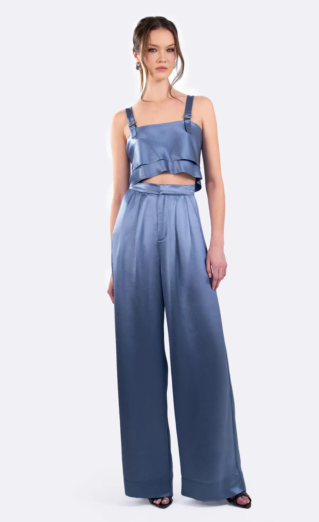 A model wearing a two-piece blue satin outfit consisting of a ruffled crop top with shoulder straps and matching high-waisted wide-leg pants, photographed against a light gray studio background.