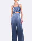 A model wearing a two-piece blue satin outfit consisting of a ruffled crop top with shoulder straps and matching high-waisted wide-leg pants, photographed against a light gray studio background.
