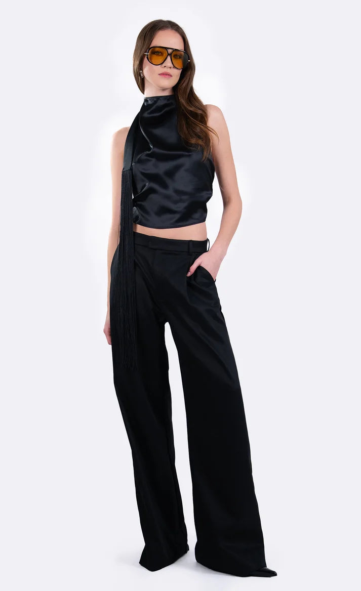 A model wearing a black satin halter crop top and matching black wide-leg trousers, styled with gold-framed sunglasses, against a plain light gray studio background.