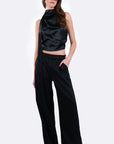 A model wearing a black satin halter crop top and matching black wide-leg trousers, styled with gold-framed sunglasses, against a plain light gray studio background.