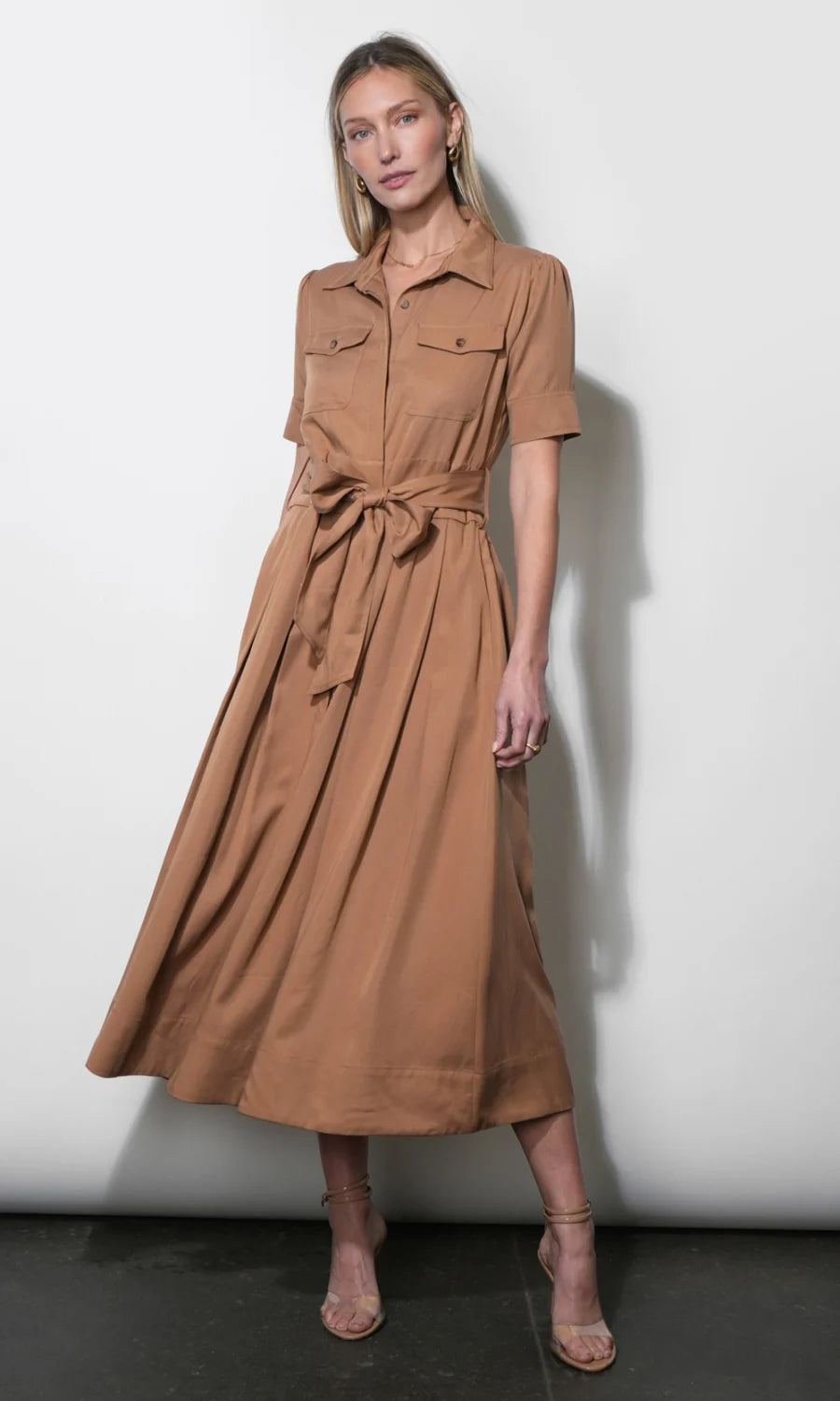 A woman modeling a tan short-sleeve midi shirt dress with a tie waist belt, standing against a plain light gray wall.