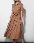 A woman modeling a tan short-sleeve midi shirt dress with a tie waist belt, standing against a plain light gray wall.