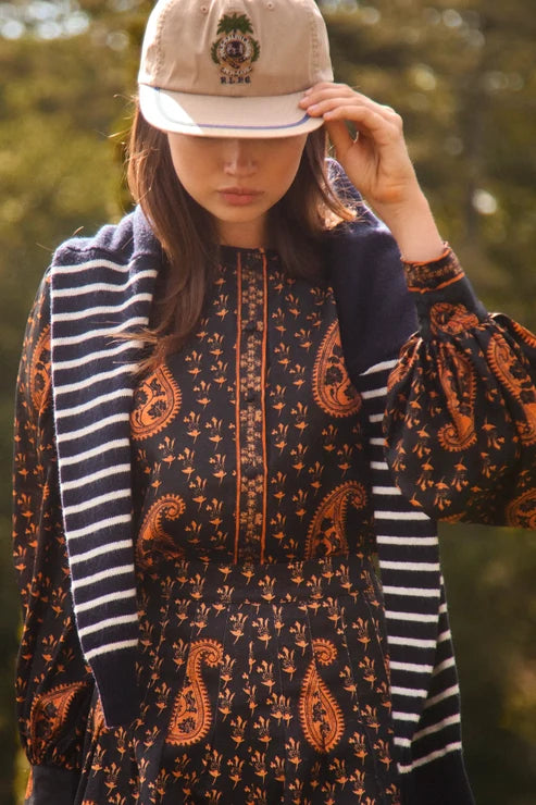 A woman outdoors wearing a beige baseball cap, a navy and orange paisley set, and a navy striped cardigan.