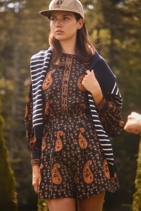 A woman wearing a dark paisley set with orange motifs, a navy and white striped cardigan, and a beige baseball cap, standing outdoors in a blurred forest setting.