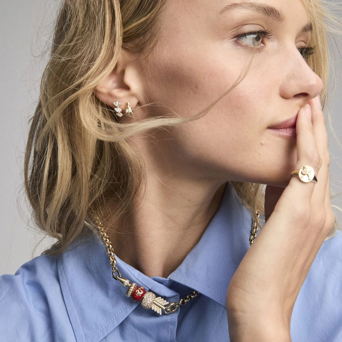 A close-up lifestyle shot of a woman wearing gold jewelry, including a gold chain necklace with a red and gold eagle pendant, a gold ring, and small gold hoop earrings, paired with a light blue collared shirt.