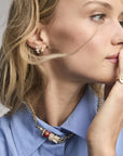 A close-up lifestyle shot of a woman wearing gold jewelry, including a gold chain necklace with a red and gold eagle pendant, a gold ring, and small gold hoop earrings, paired with a light blue collared shirt.