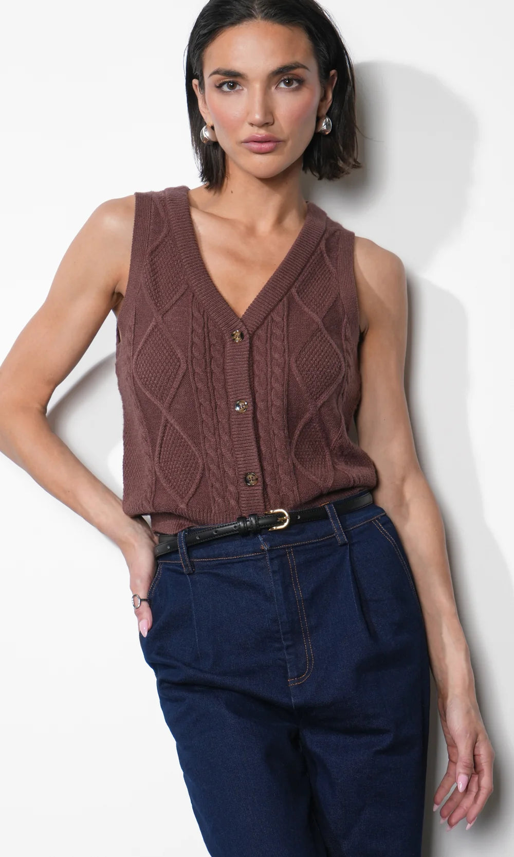A woman modeling a sleeveless brown cable-knit vest with a V-neck and button front, paired with dark blue jeans and a black belt, against a plain white wall.