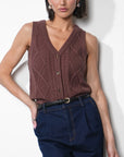 A woman modeling a sleeveless brown cable-knit vest with a V-neck and button front, paired with dark blue jeans and a black belt, against a plain white wall.