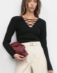 Woman wearing a black long-sleeve top with criss-cross design and beige pants, holding a red clutch against a white background.