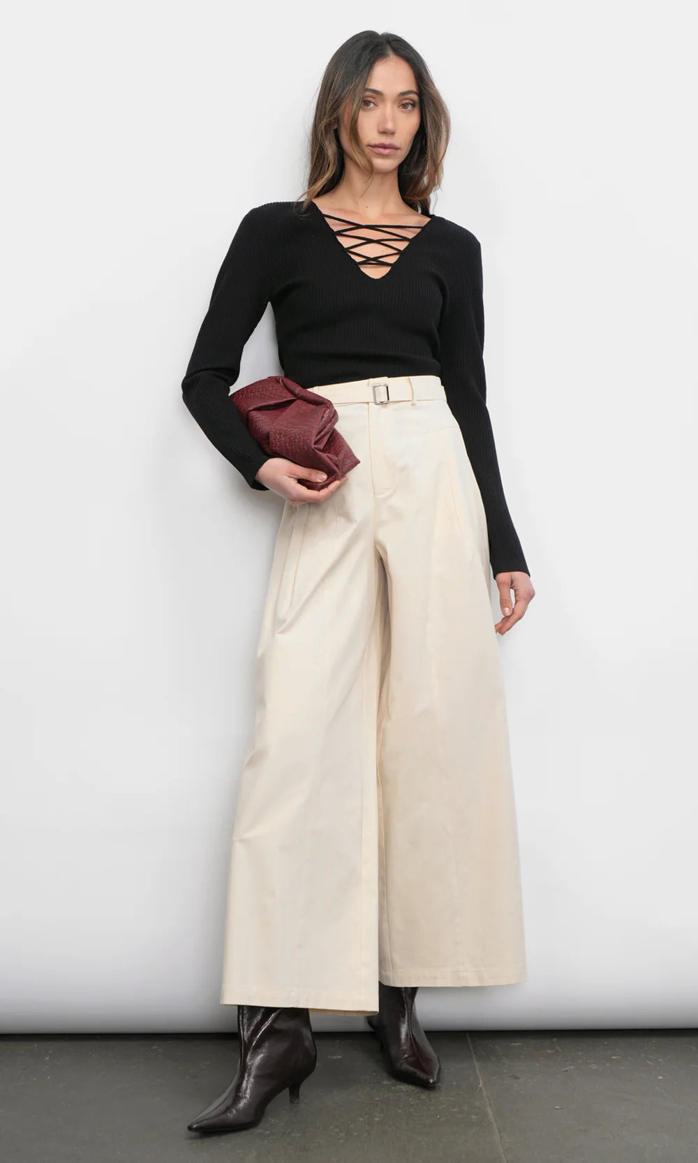 Woman wearing a black top and beige wide-leg pants holding a red clutch against a white background, full body