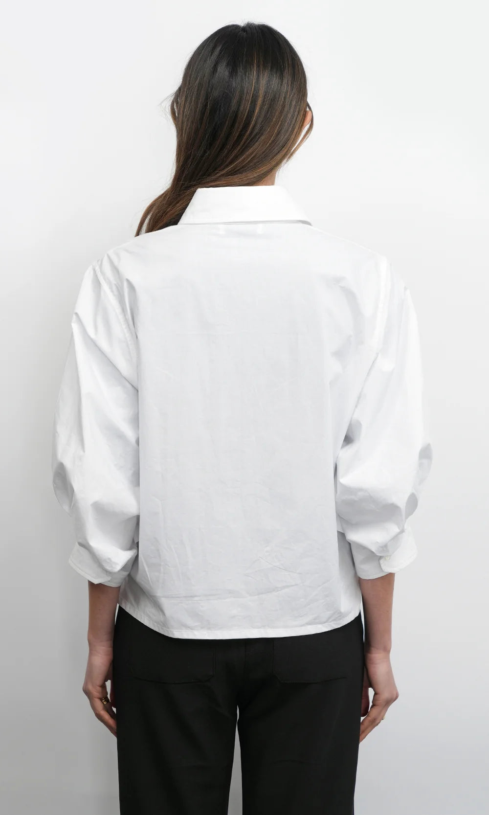 A woman viewed from the back wearing a white long-sleeve button-up shirt with voluminous sleeves and black trousers, set against a plain white studio background.