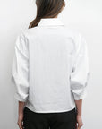 A woman viewed from the back wearing a white long-sleeve button-up shirt with voluminous sleeves and black trousers, set against a plain white studio background.