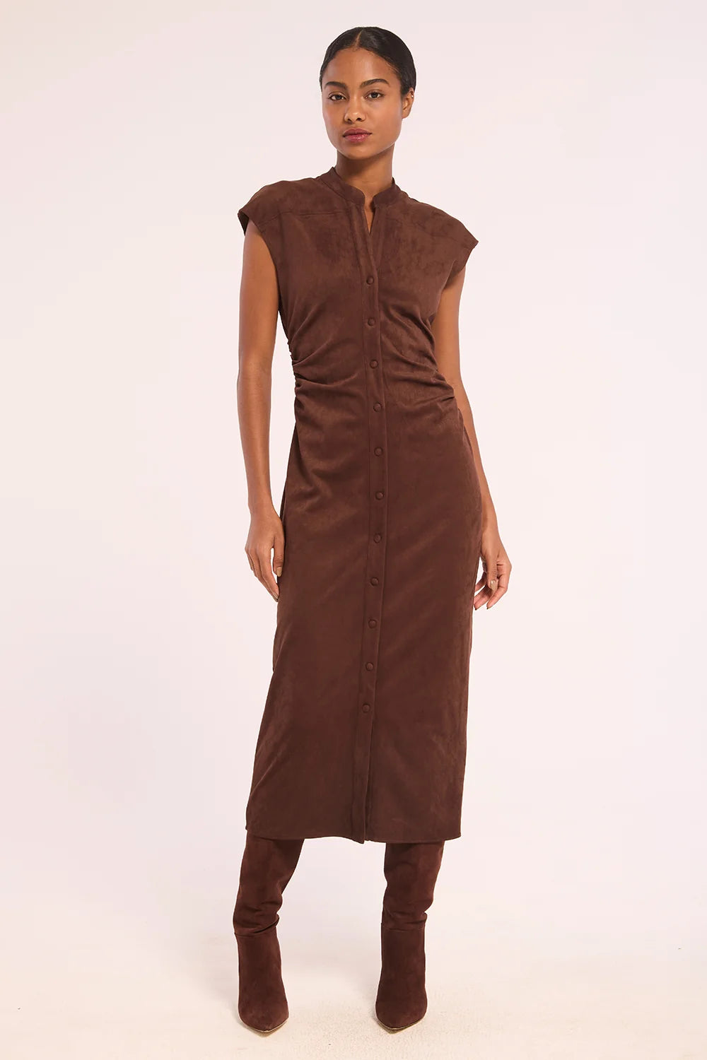 A model wearing a brown sleeveless midi dress with a button-down front and gathered waist, paired with matching brown boots, against a plain off-white background.