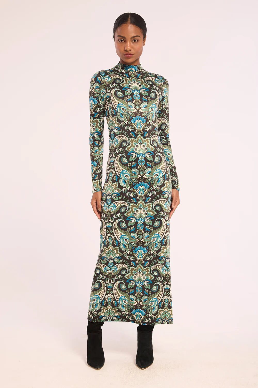 A woman modeling a long-sleeve, high-neck maxi dress with an intricate paisley pattern in black, teal, blue, and beige, paired with black ankle boots against a solid light background.