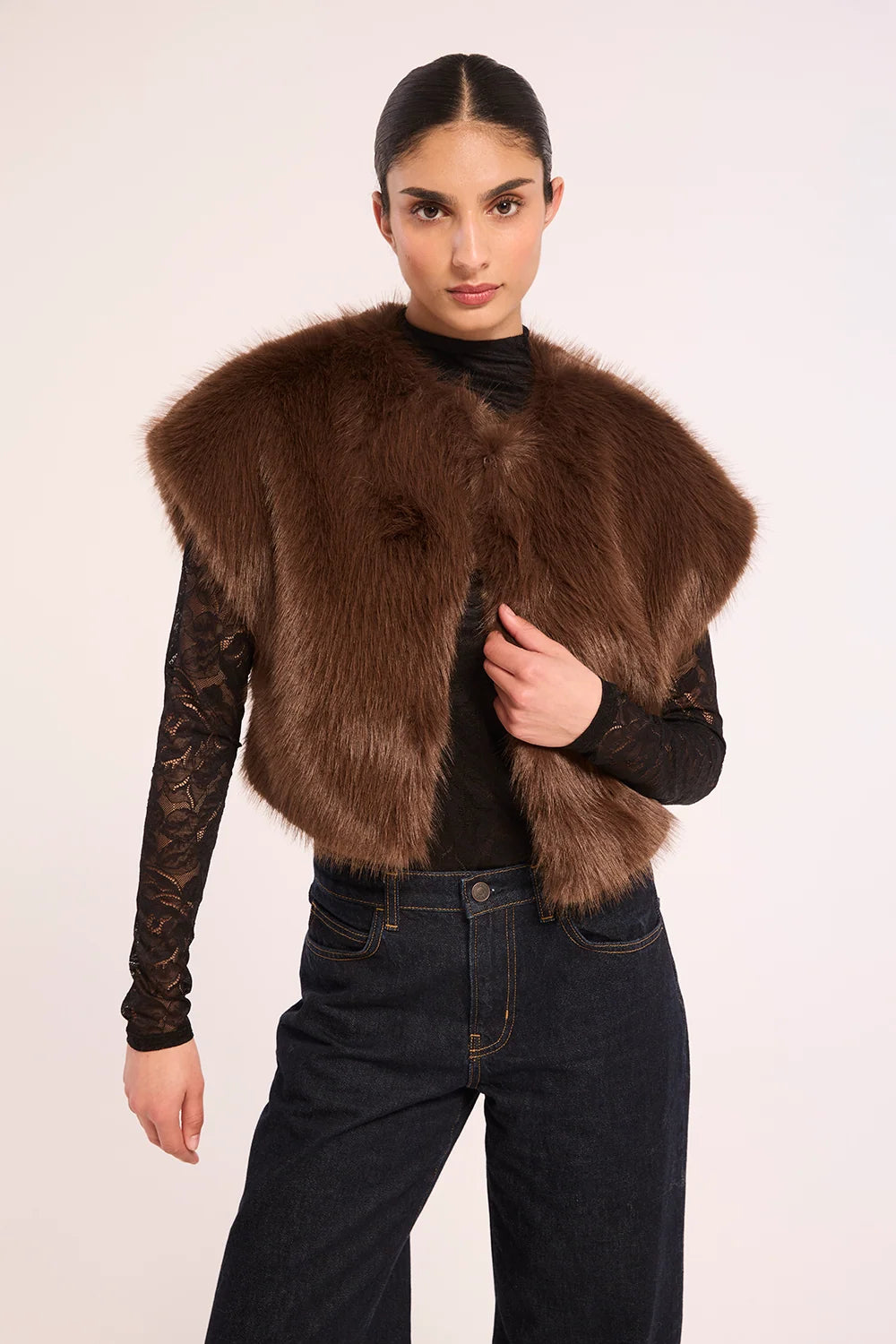 A model wearing a brown faux fur vest over a black lace long-sleeve top and dark blue jeans, photographed against a plain light background.