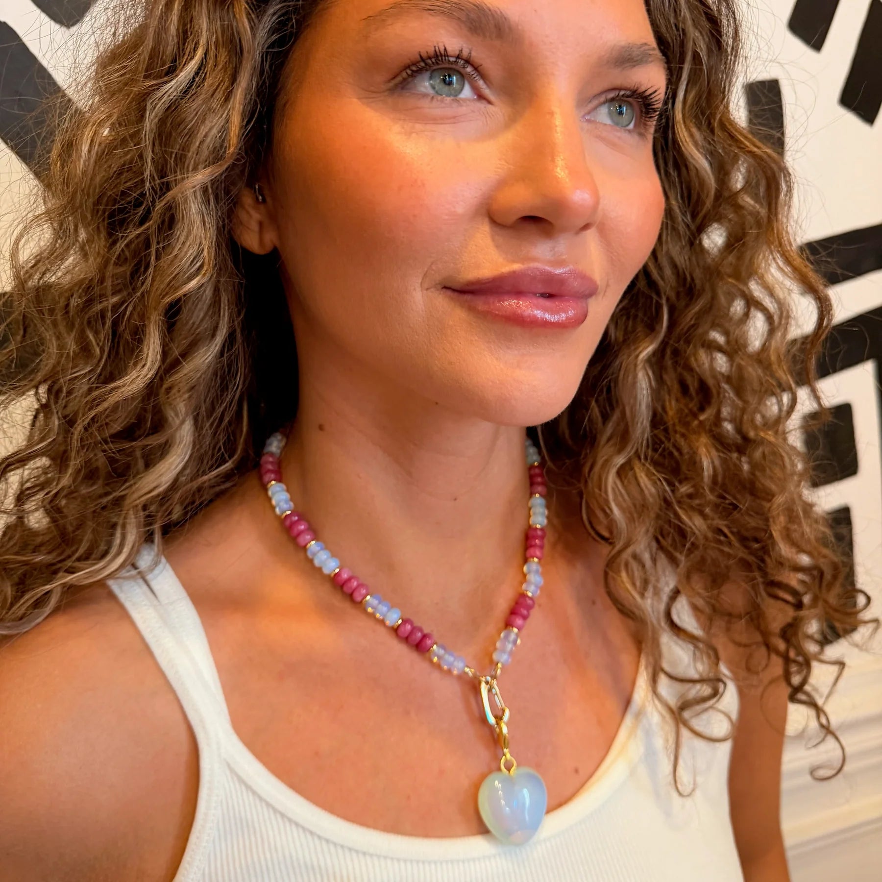 Woman wearing a beaded necklace with a heart pendant in front of a geometric patterned wall.