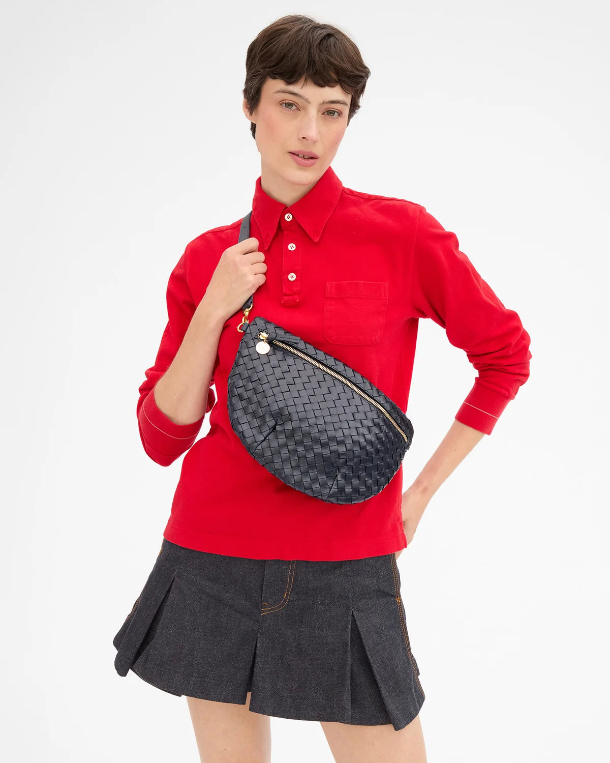 A model wearing a red long-sleeve button-up shirt and a dark gray pleated mini skirt, carrying a dark navy woven leather fanny pack across their waist