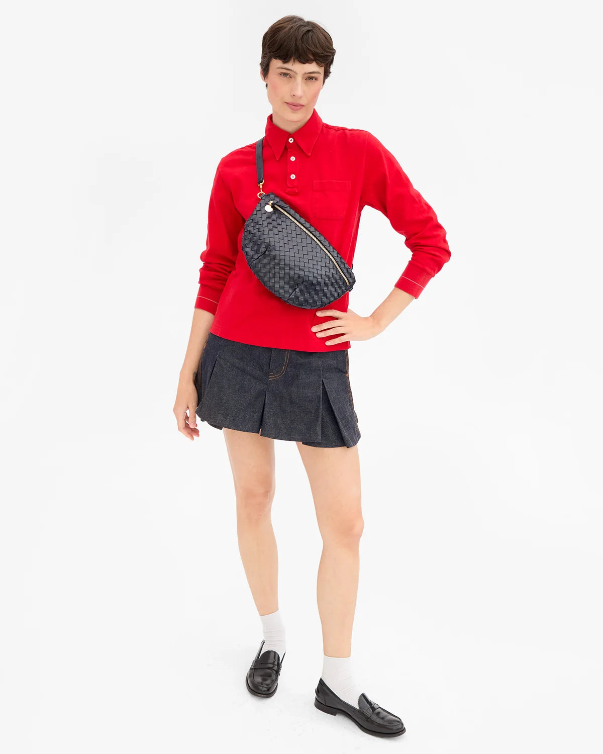A model wearing a vibrant red long-sleeve polo shirt, a dark gray pleated mini skirt, white socks, and black loafers, accessorized with a dark navy woven crossbody bag.